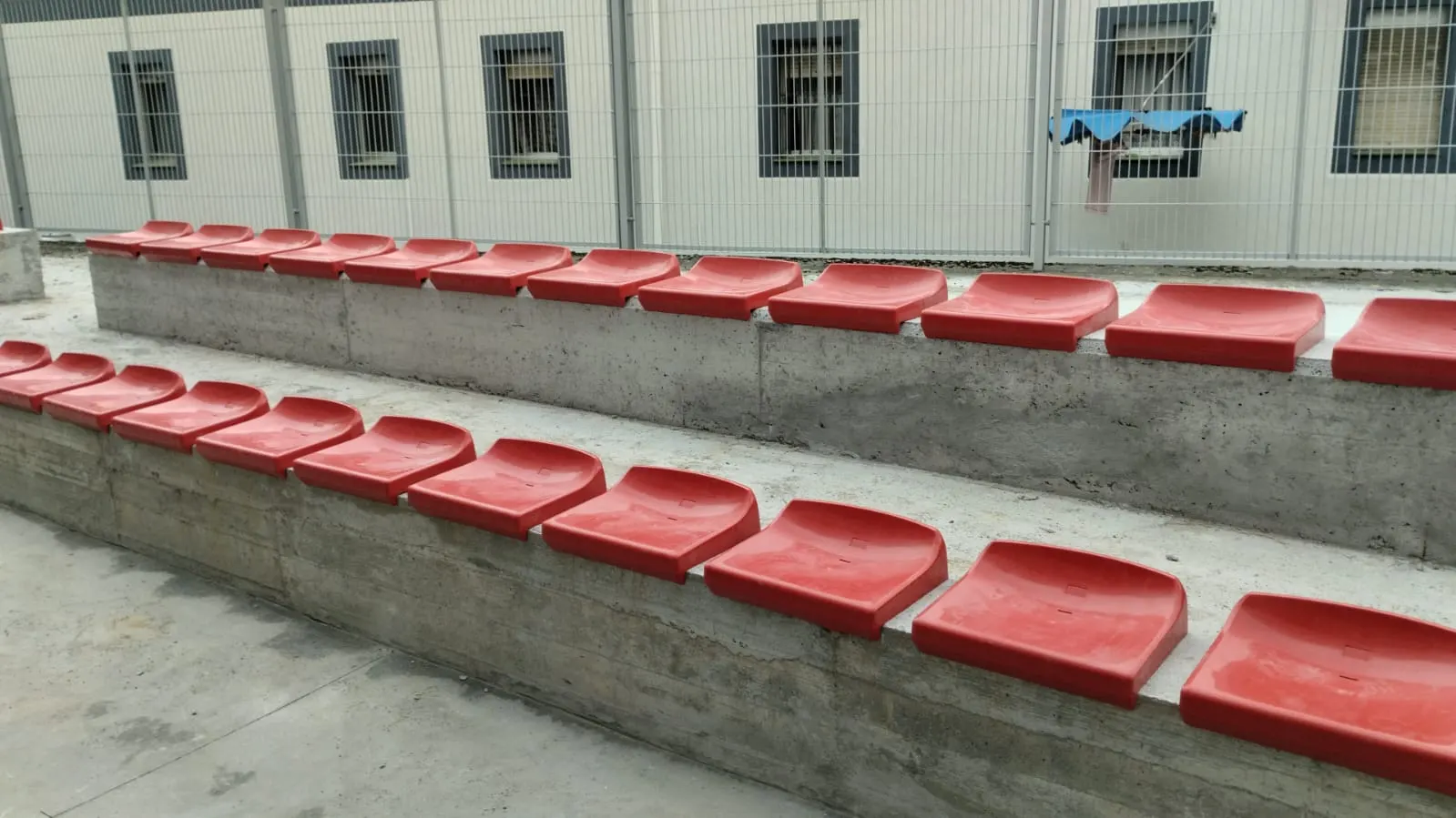 Grada de hormigón con dos filas de asientos rojos de plástico en una instalación deportiva, junto a una valla metálica