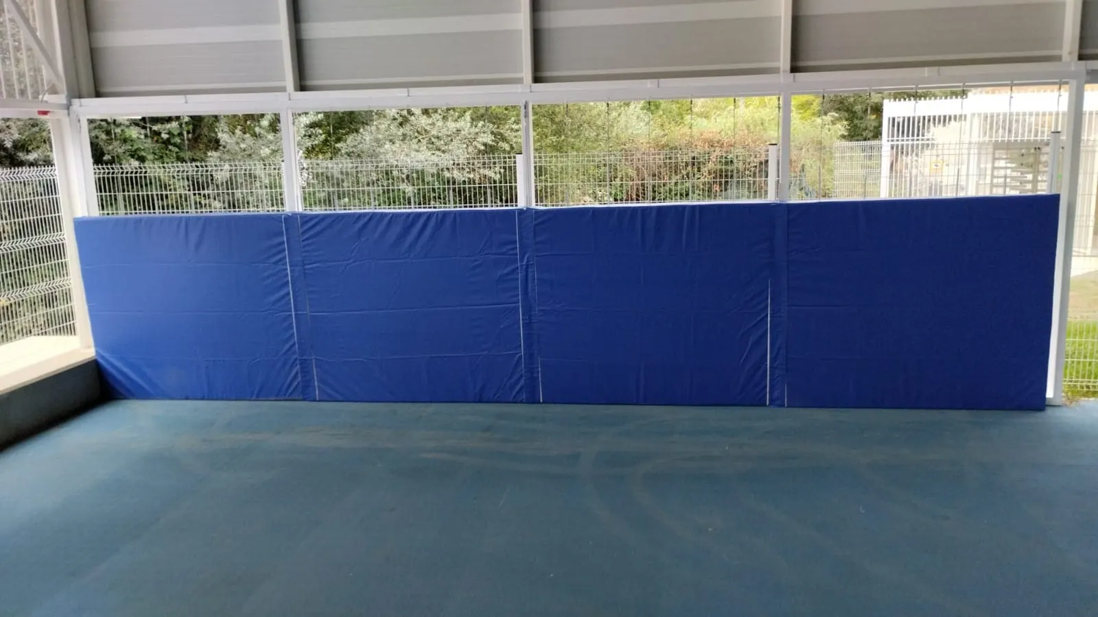 Protecciones acolchadas azules en la pared de una pista cubierta de una instalación deportiva, con valla metálica detrás