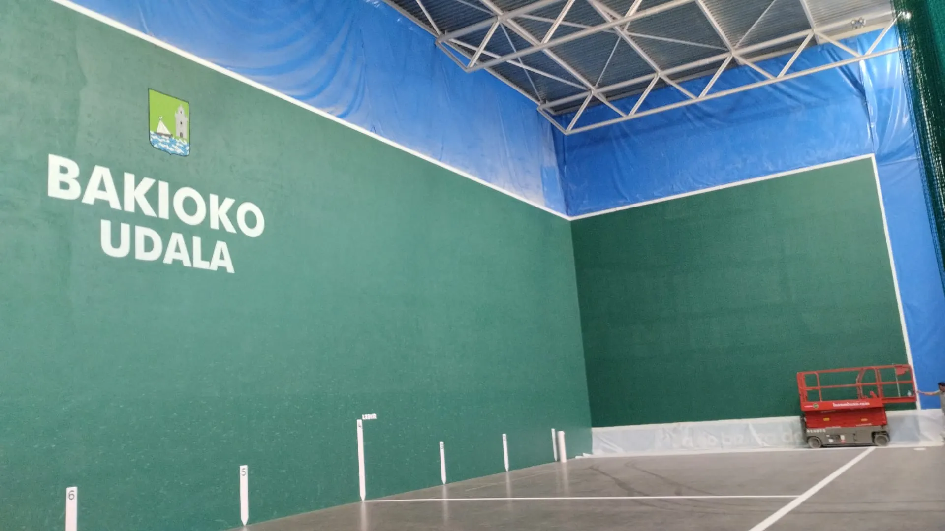 Frontón cubierto de Bakio con paredes verdes recién pintadas, rótulo «Bakioko Udala» y una plataforma elevadora roja al fondo