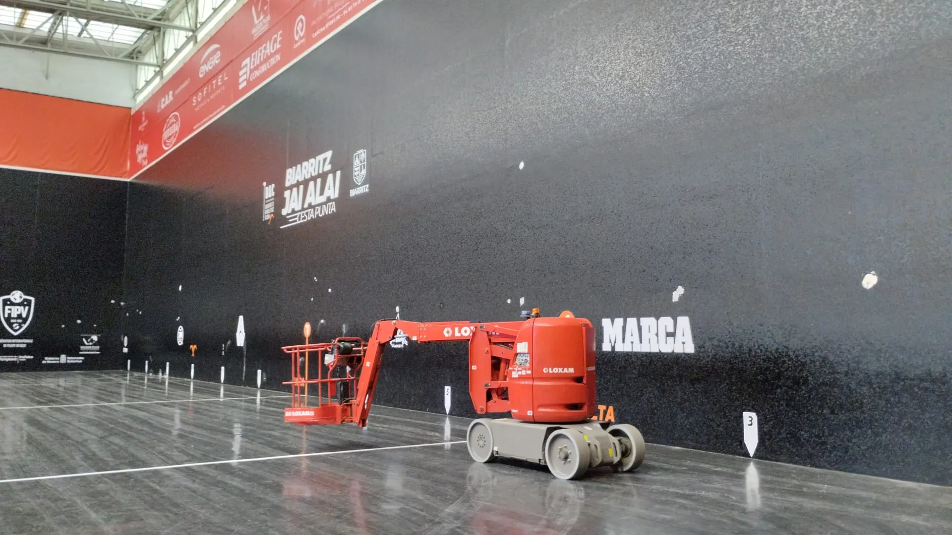 Plataforma elevadora roja de Loxam sobre la cancha de un frontón de cesta punta con paredes negras en Biarritz