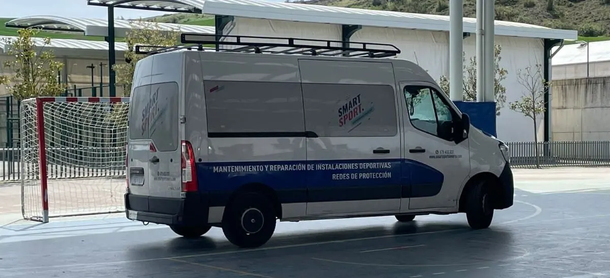 Furgoneta blanca de Smart Sport con baca, aparcada en una pista polideportiva cubierta junto a una portería de fútbol sala
