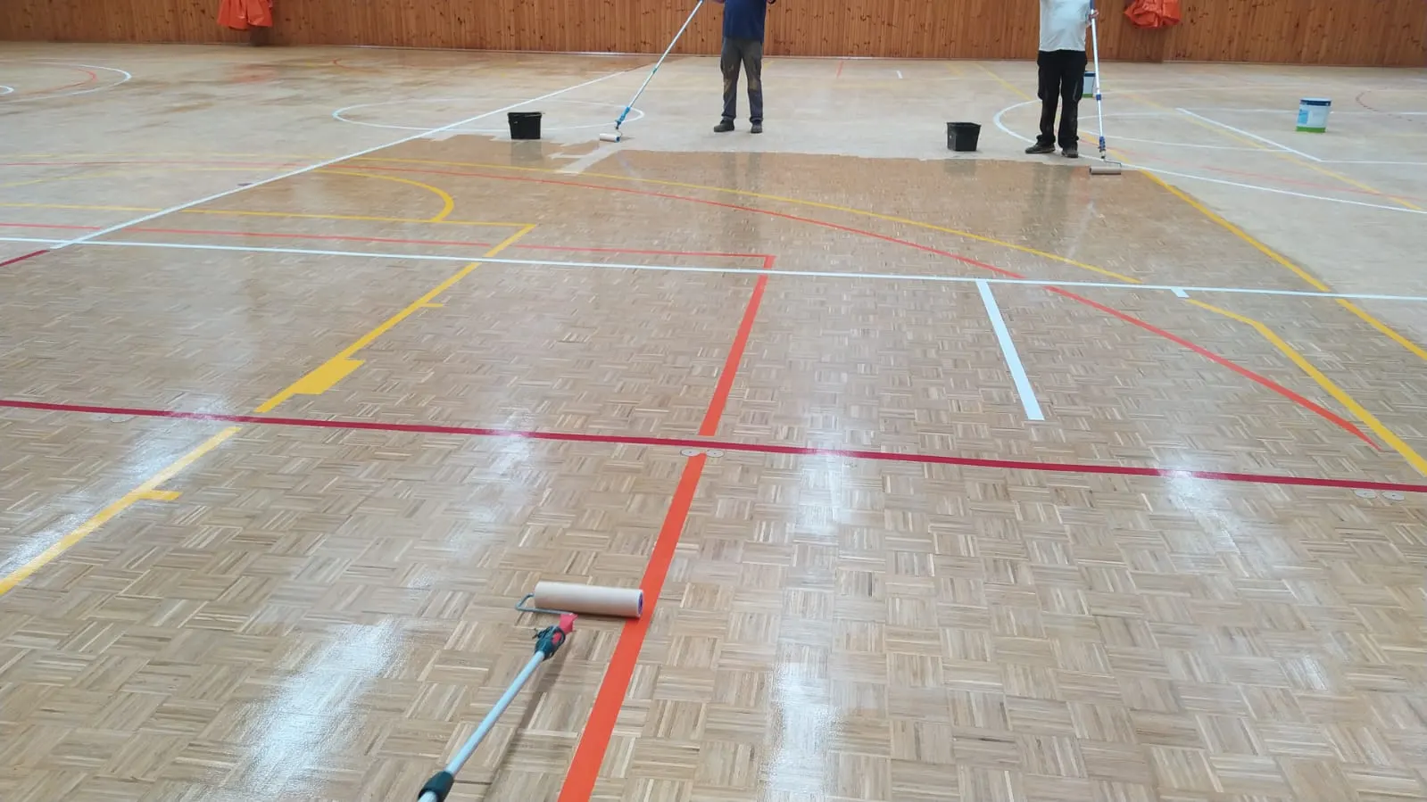 Dos operarios barnizan con rodillo el parquet de una pista deportiva interior con líneas de marcaje de colores