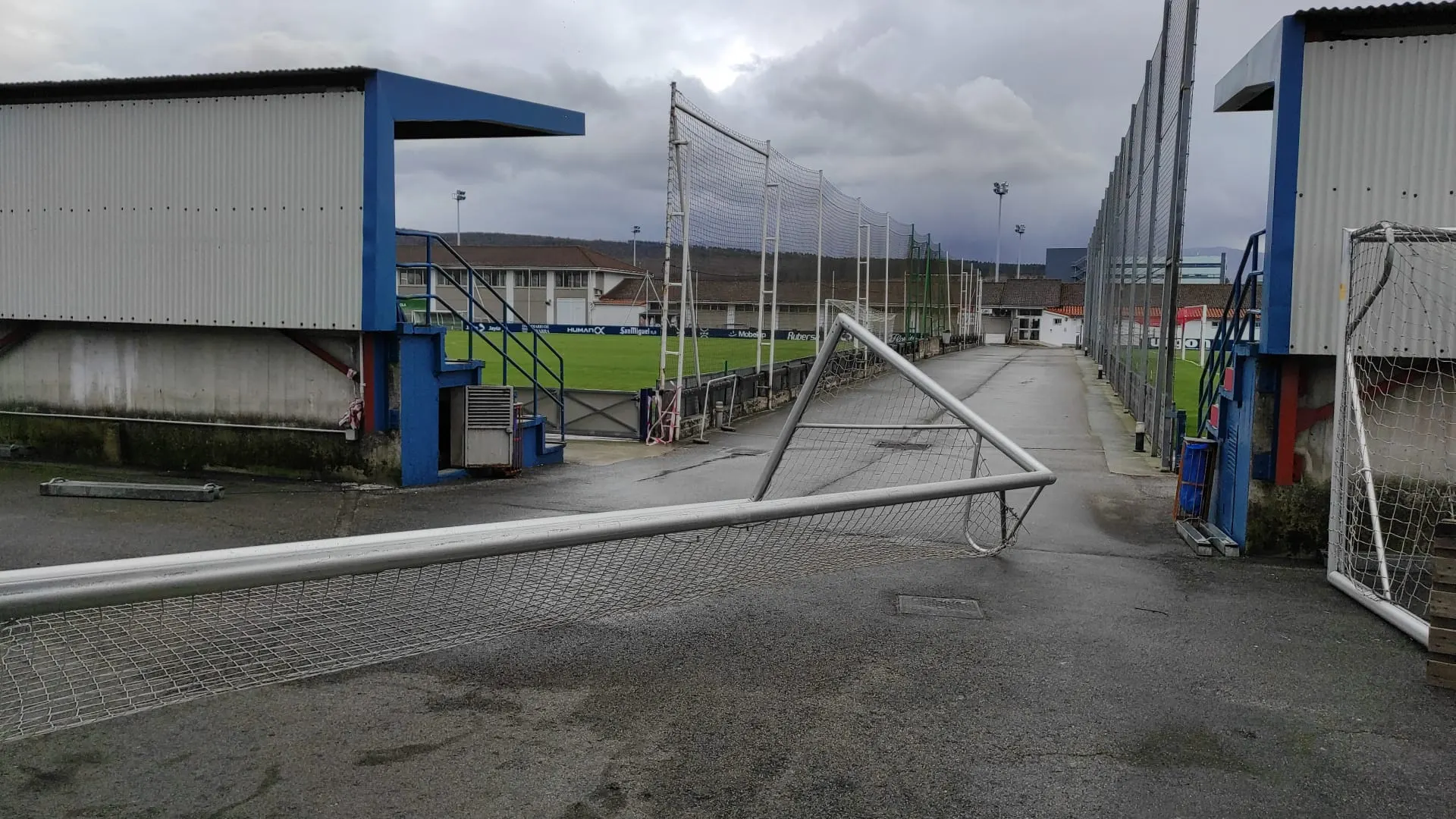 Portería de fútbol caída sobre el asfalto mojado entre gradas azules de unas instalaciones deportivas con campos de césped