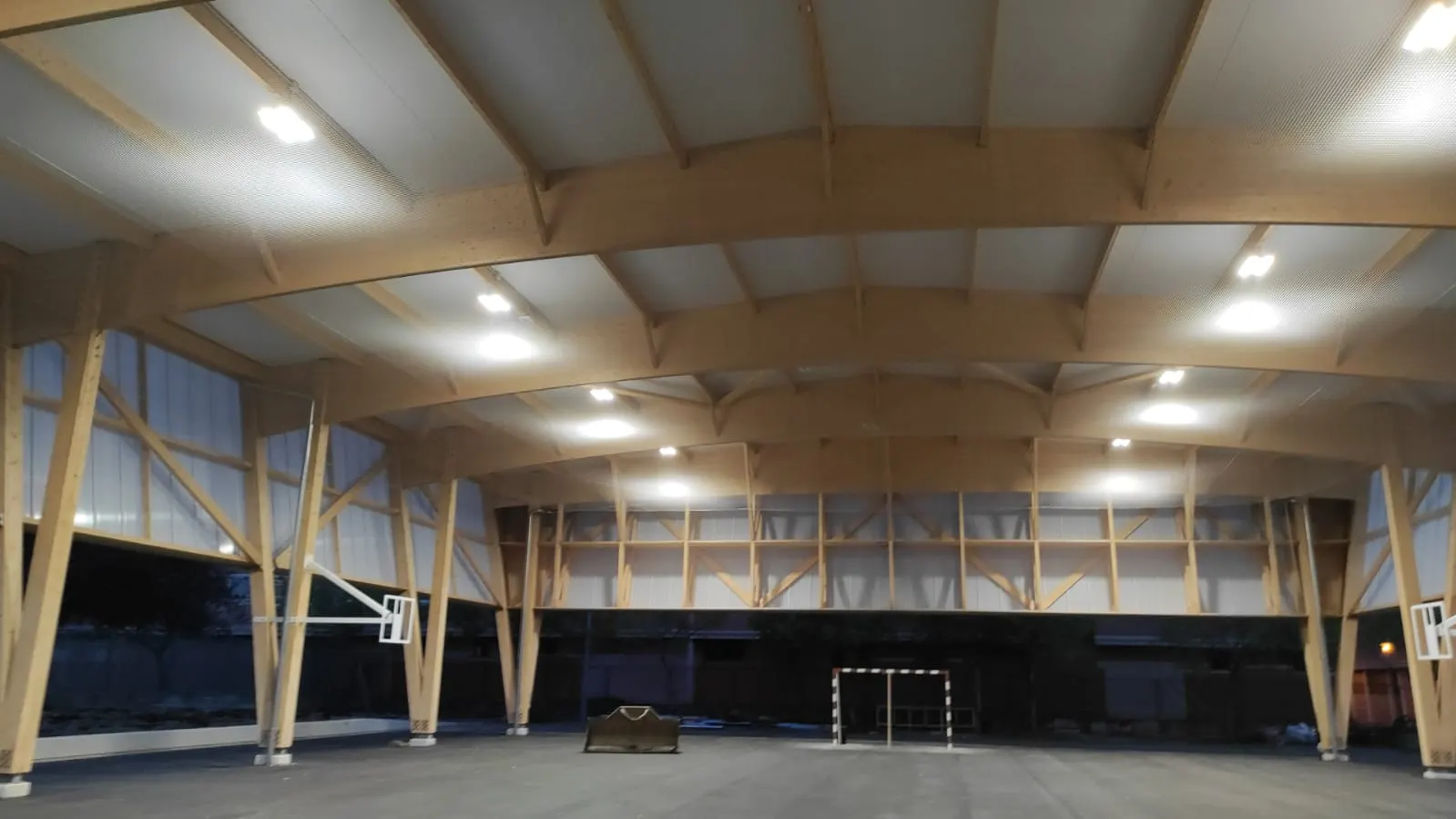Pista polideportiva cubierta con arcos de madera laminada, canastas laterales y portería de fútbol sala al fondo