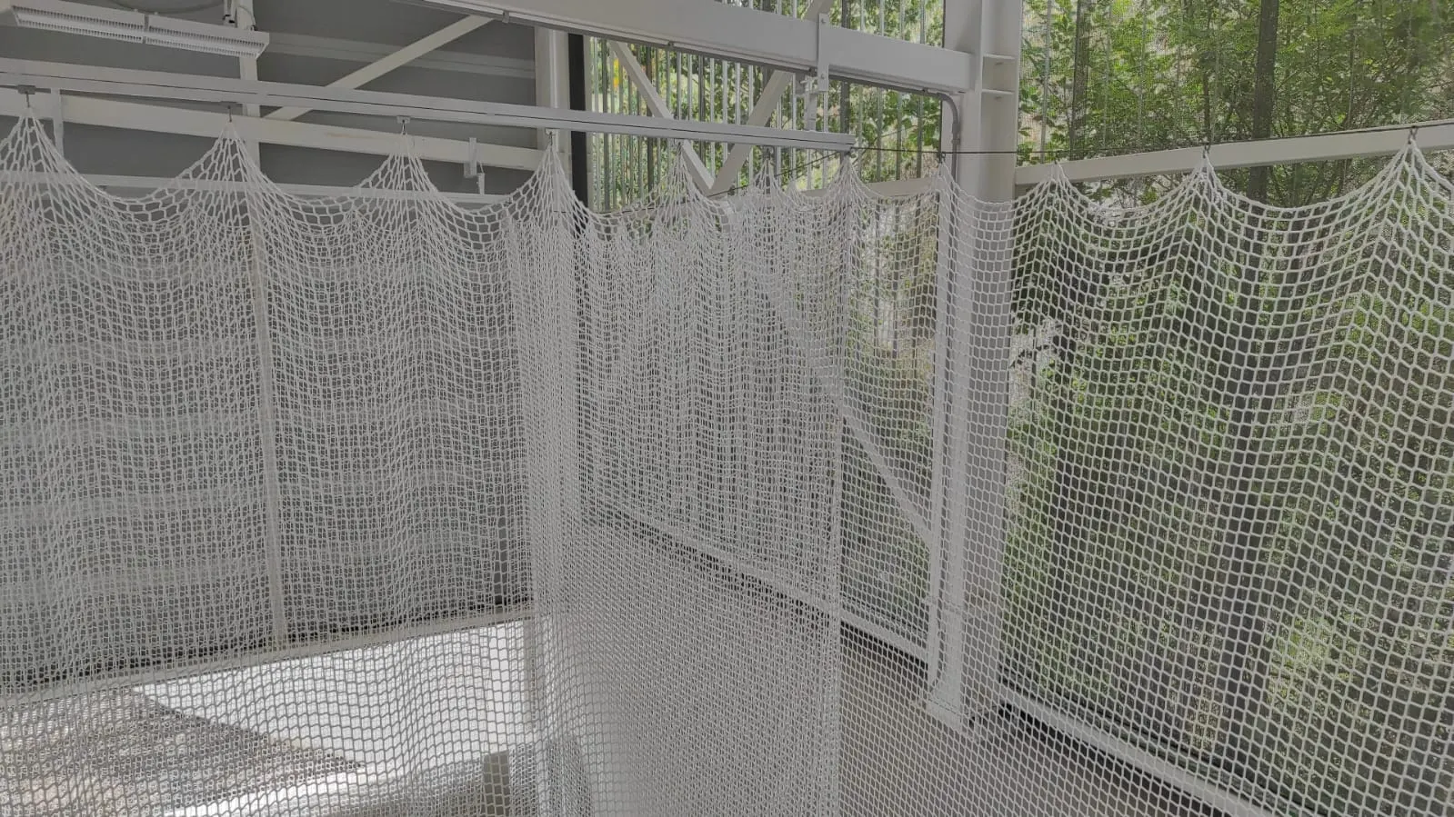 Red blanca de protección colgada de una estructura metálica que cierra una instalación deportiva junto a una zona arbolada