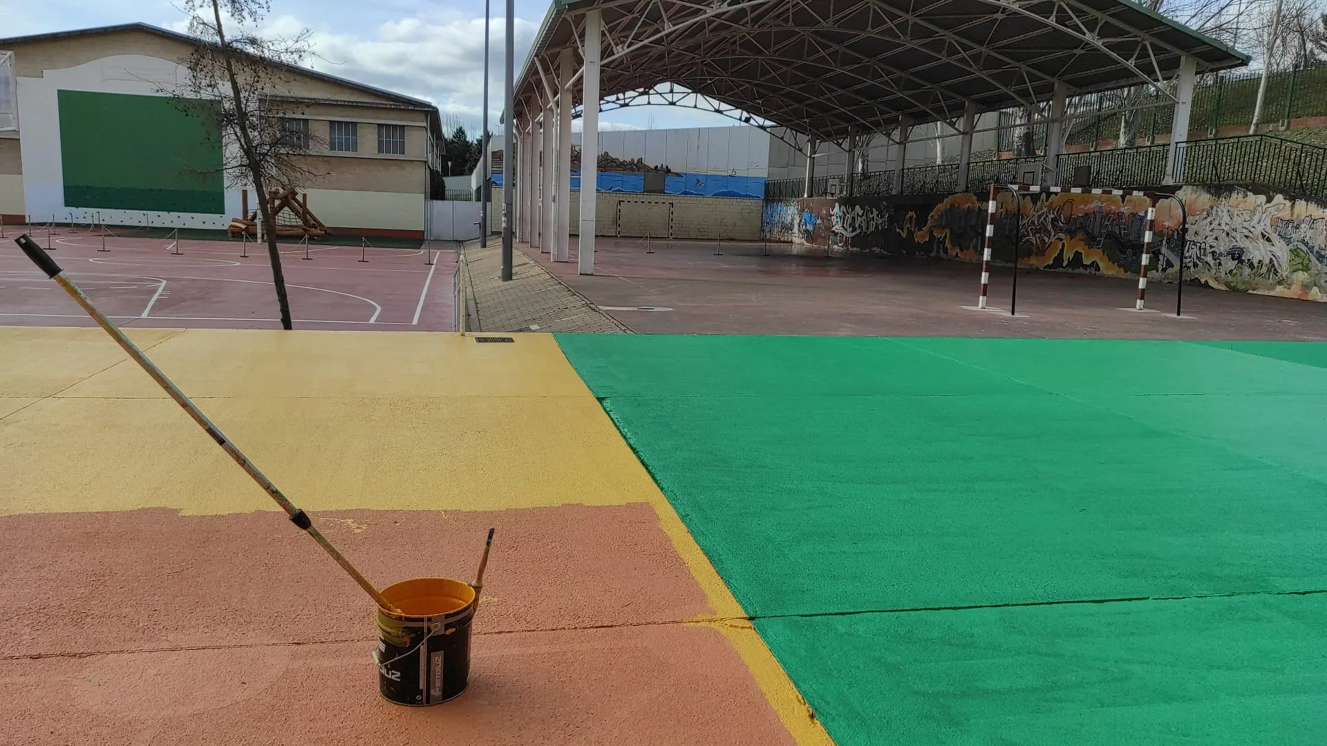 Pista deportiva recién pintada de verde, amarillo y rojo, con bote de pintura y rodillo junto a porterías bajo cubierta