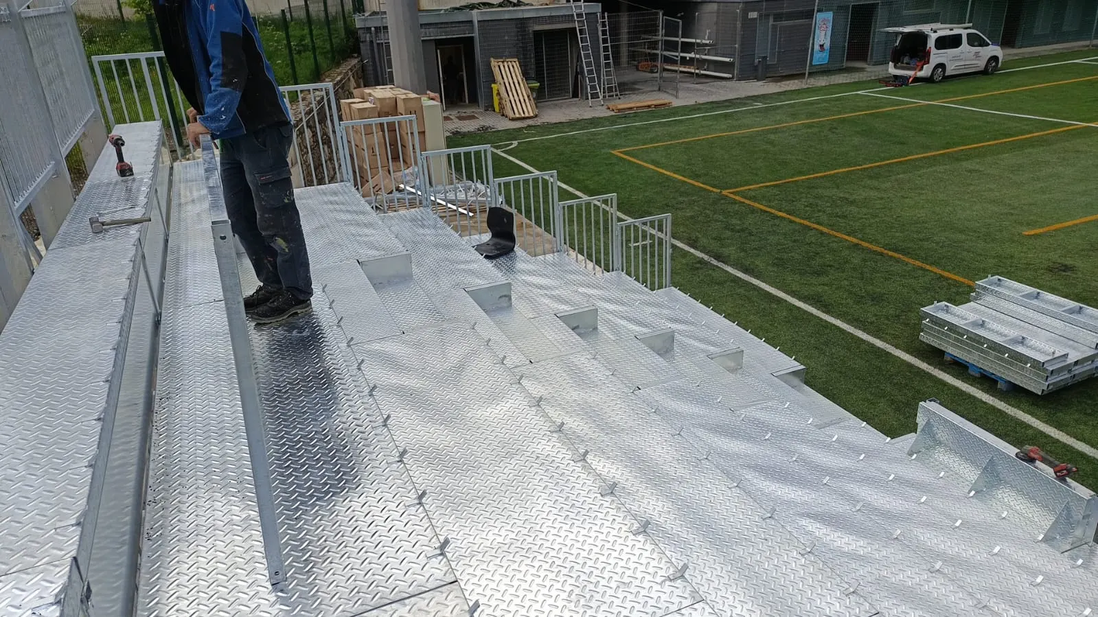 Operario montando una grada de chapa de aluminio estriada junto a un campo de fútbol de césped artificial