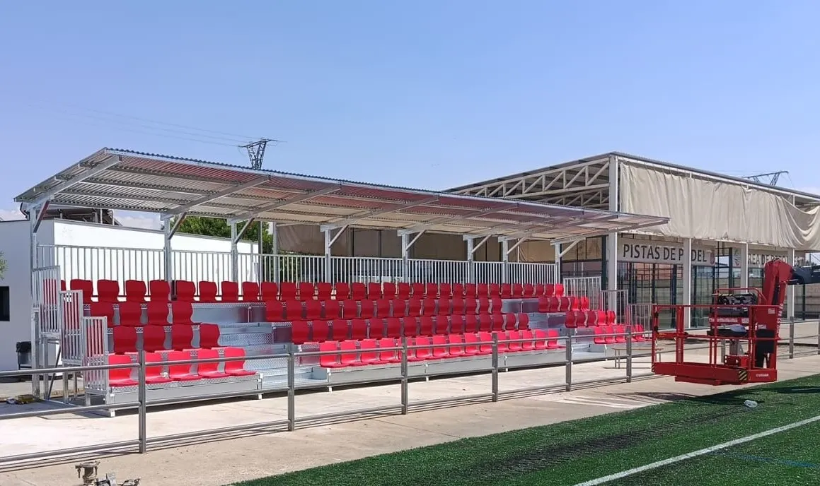 Montaje de grada metálica cubierta con asientos rojos junto a un campo de césped artificial, con plataforma elevadora