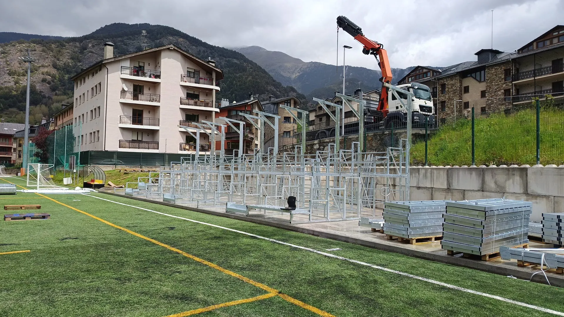 Montaje de una grada de estructura metálica junto a un campo de césped artificial, con un camión grúa y montañas al fondo