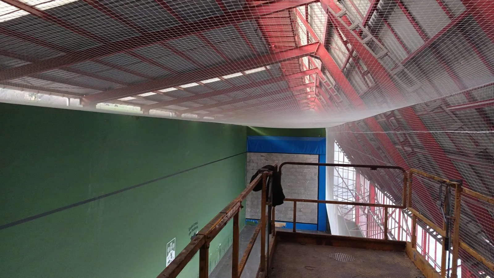 Red de protección blanca instalada bajo la cubierta metálica roja de un frontón, vista desde una plataforma elevadora