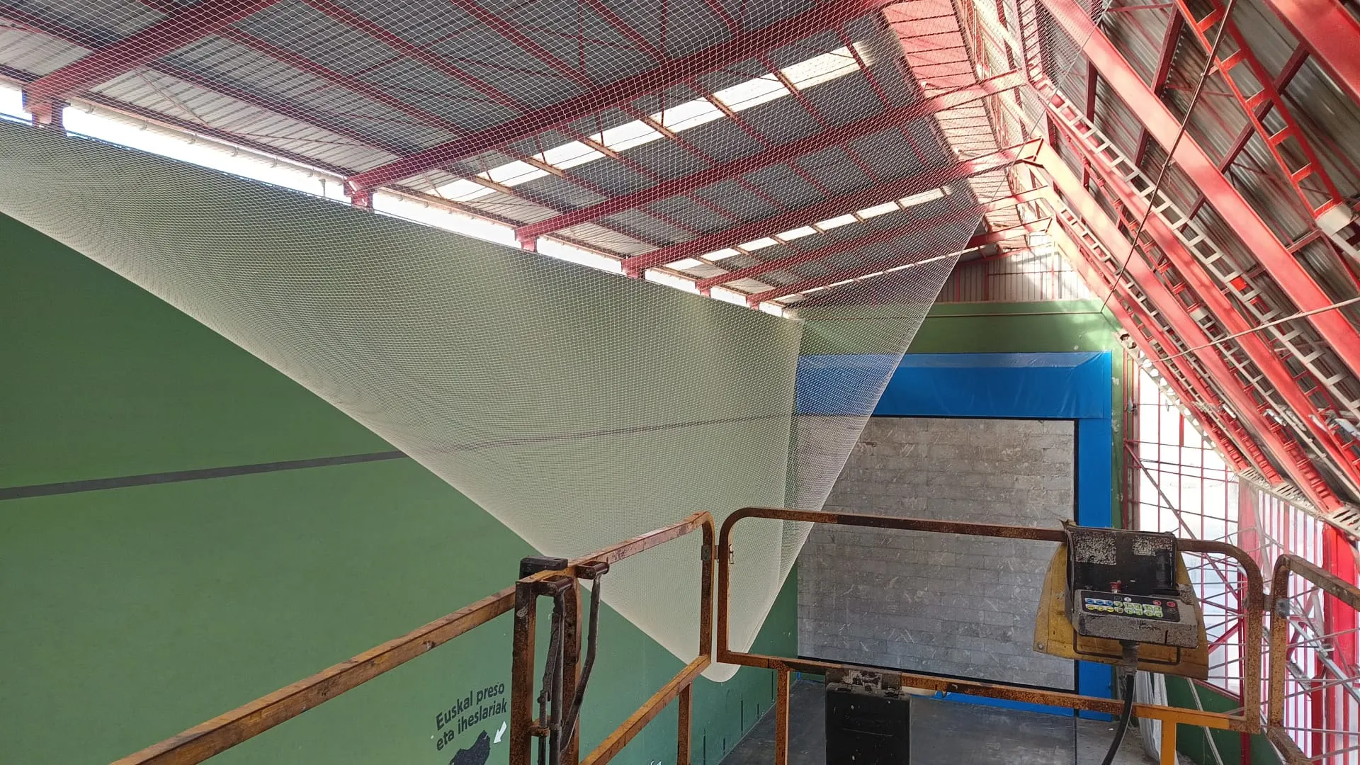 Red de protección blanca instalada bajo la cubierta metálica roja de un frontón, vista desde una plataforma elevadora