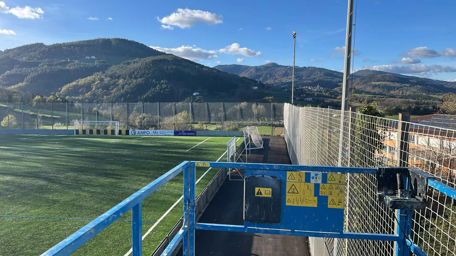 Red perimetral alta junto a un campo de fútbol de césped artificial, con una barandilla azul y montes al fondo