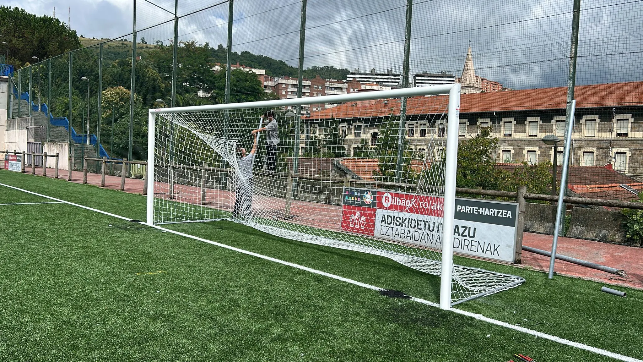 Dos operarios colocan la red en una portería de fútbol blanca sobre un campo de césped artificial, en plena instalación