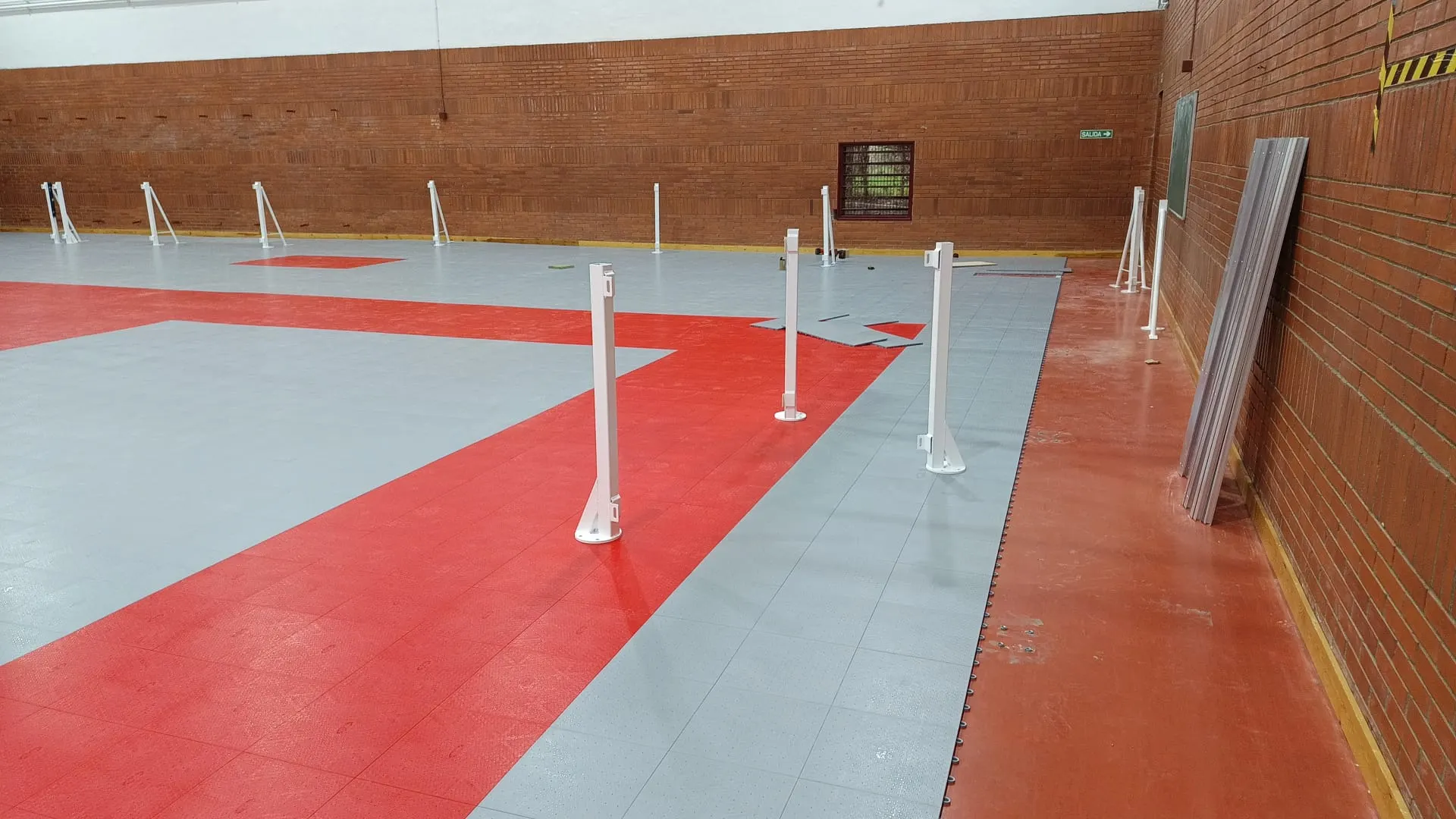 Pista deportiva cubierta con suelo modular gris y rojo, postes blancos en instalación y paredes de ladrillo visto