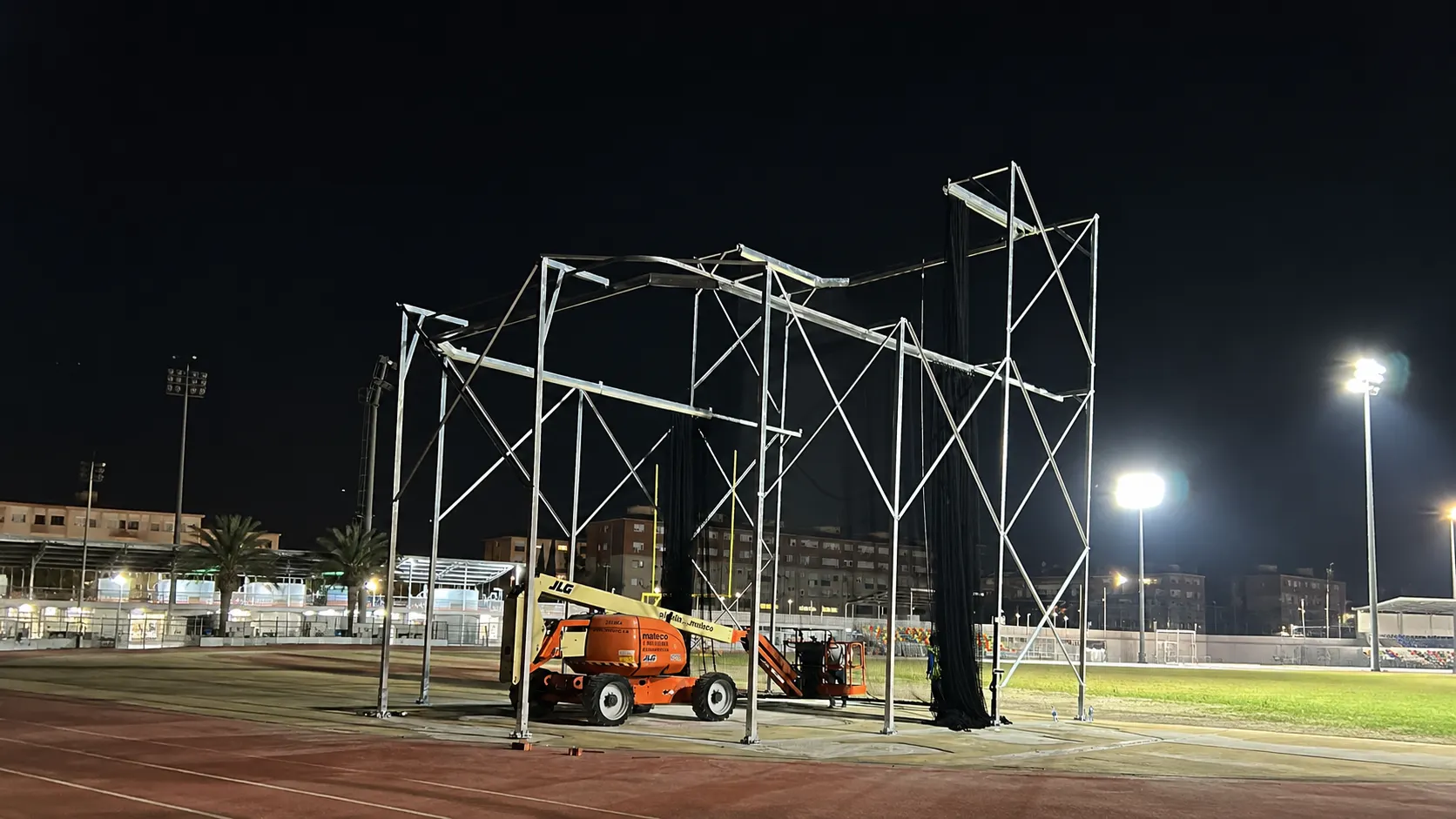Montaje nocturno de una jaula de bateo con estructura metálica y redes negras junto a una plataforma elevadora naranja